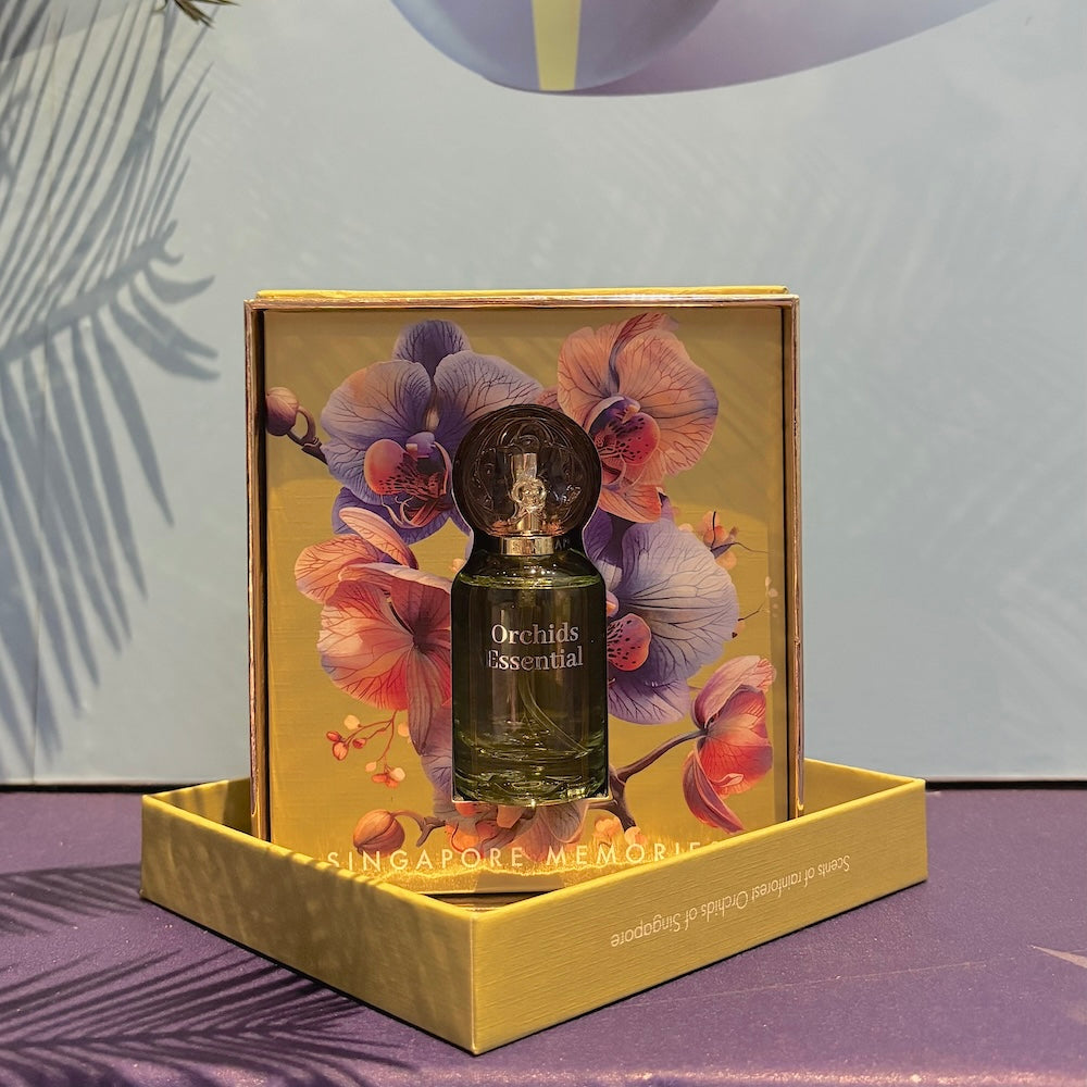 A bottle of Orchids Essential perfume presented in a yellow box with a colorful floral design. The box is displayed on a surface with a shadow of a plant leaf.