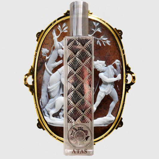 A bottle of Atas fragrance placed in front of an ornate, vintage-style frame depicting classical figures. The bottle has a decorative pattern and the brand name 'Atas' is visible.