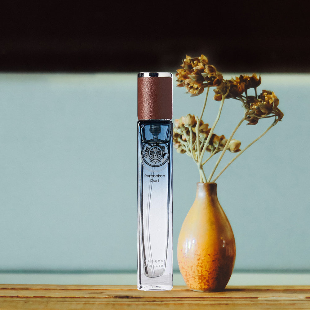 A bottle of Peranakan Oud perfume placed next to a vase with dried flowers, on a wooden surface with a softly focused background.