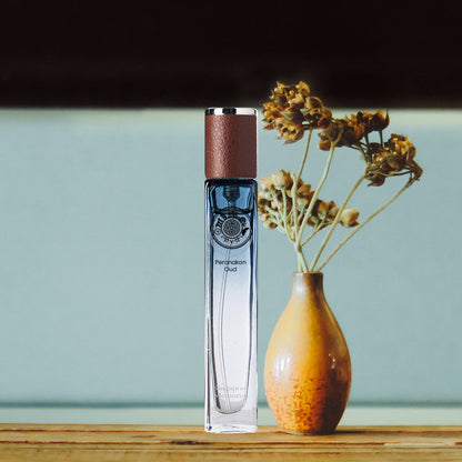 A bottle of Peranakan Oud perfume placed next to a vase with dried flowers, on a wooden surface with a softly focused background.