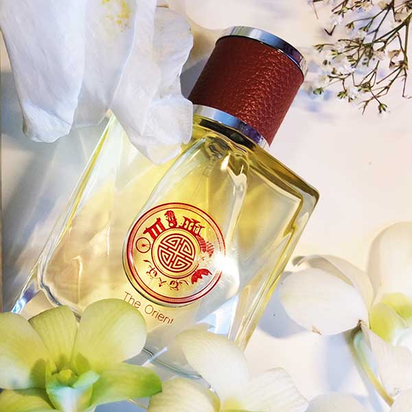 Perfume bottle with a red cap and visible brand logo  'the orient' by singapore memories, surrounded by flowers.