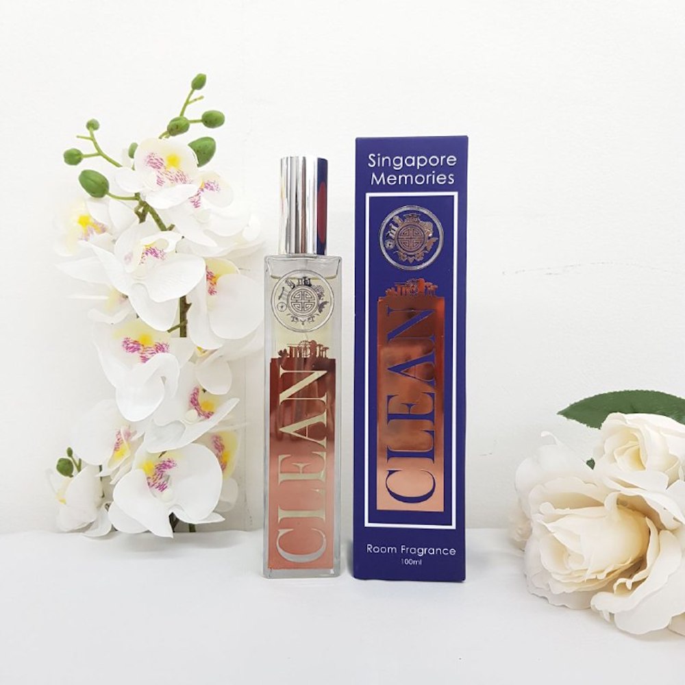 CLEAN room fragrance by singapore memories in a glass and metal bottle next to a blue gift box, in front of white background, surrounded by white orchids and rose