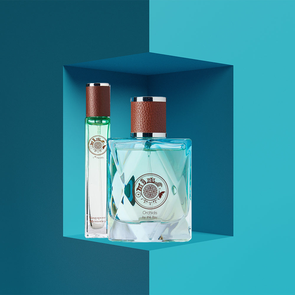 Two bottles of perfume, with the first one showing a green cap and the second one a brown cap. Both display the label 'Orchids by the Bay' and are set against a blue geometric pattern background.