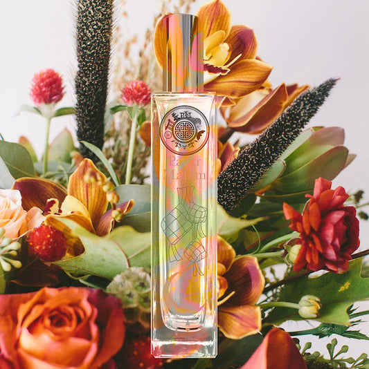 A clear glass bottle of fragrance with a floral design, positioned in a vase with various colorful flowers and decorative elements.