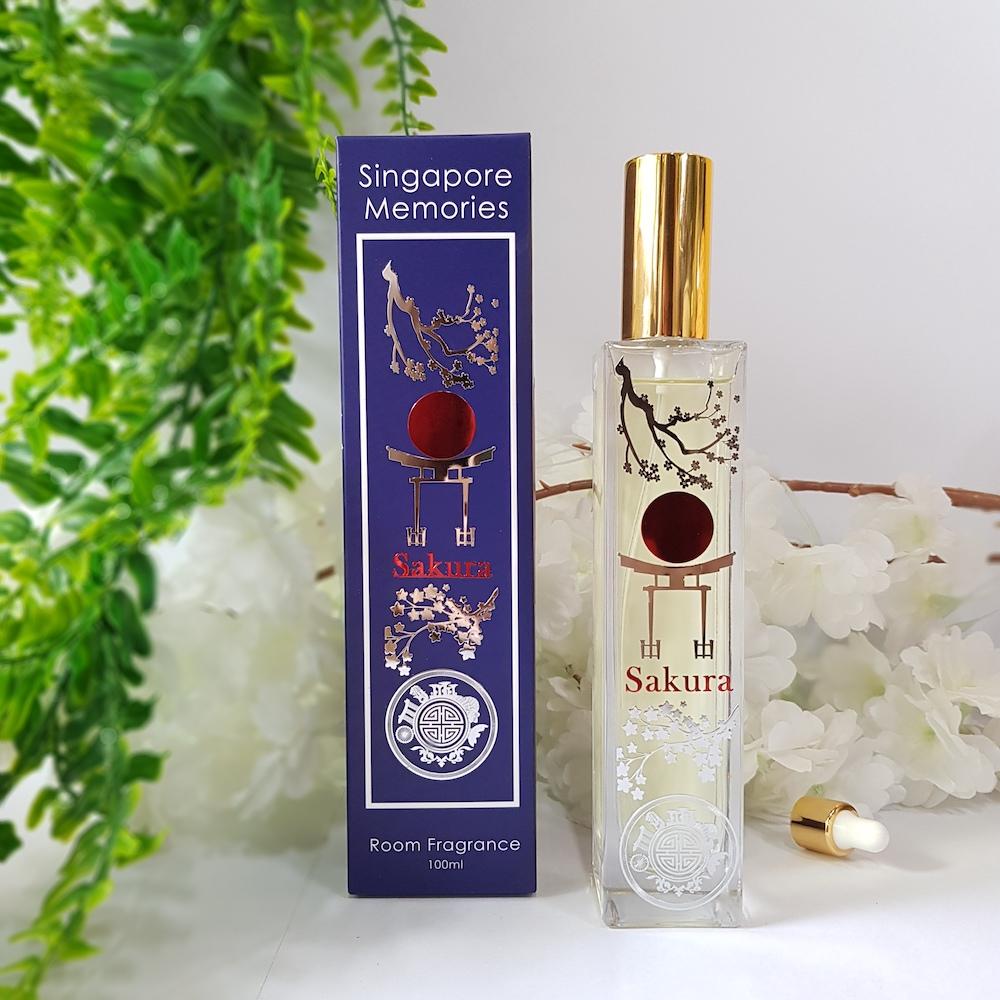 Sakura perfume glass bottle with elegant design, next to a blue gift box, in front of white flowers and next to green fern leaves