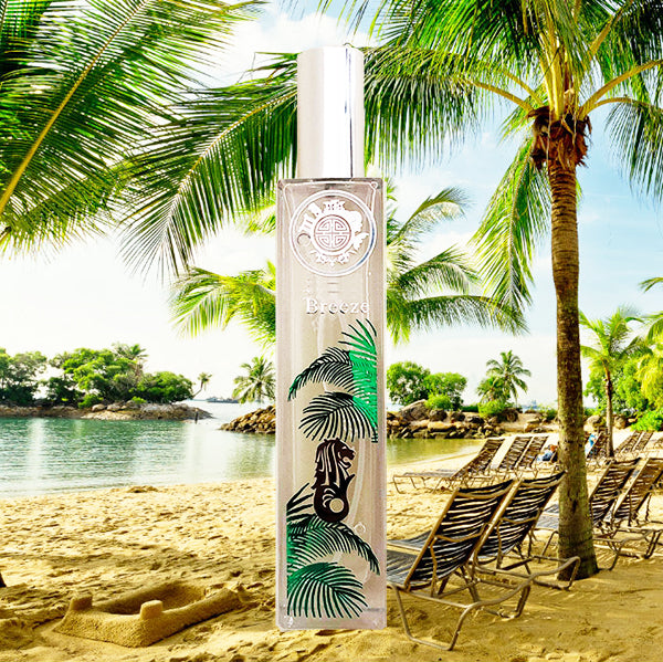 A tall, clear bottle of perfume with a label featuring a black and green singapore breeze design, placed on a sandy beach with palm trees in the background.