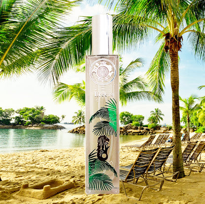 A tall, clear bottle of perfume with a label featuring a black and green singapore breeze design, placed on a sandy beach with palm trees in the background.
